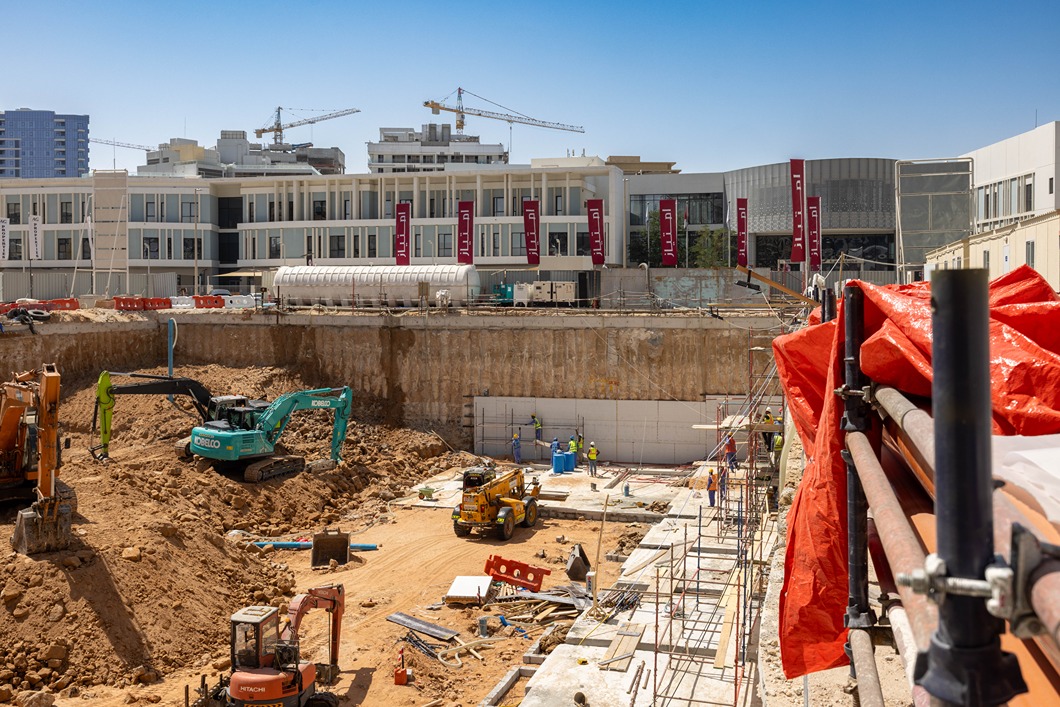 Cove Edition I By Imtiaz 1 A construction site with heavy machinery, workers, and a partially dug foundation next to a modern building.